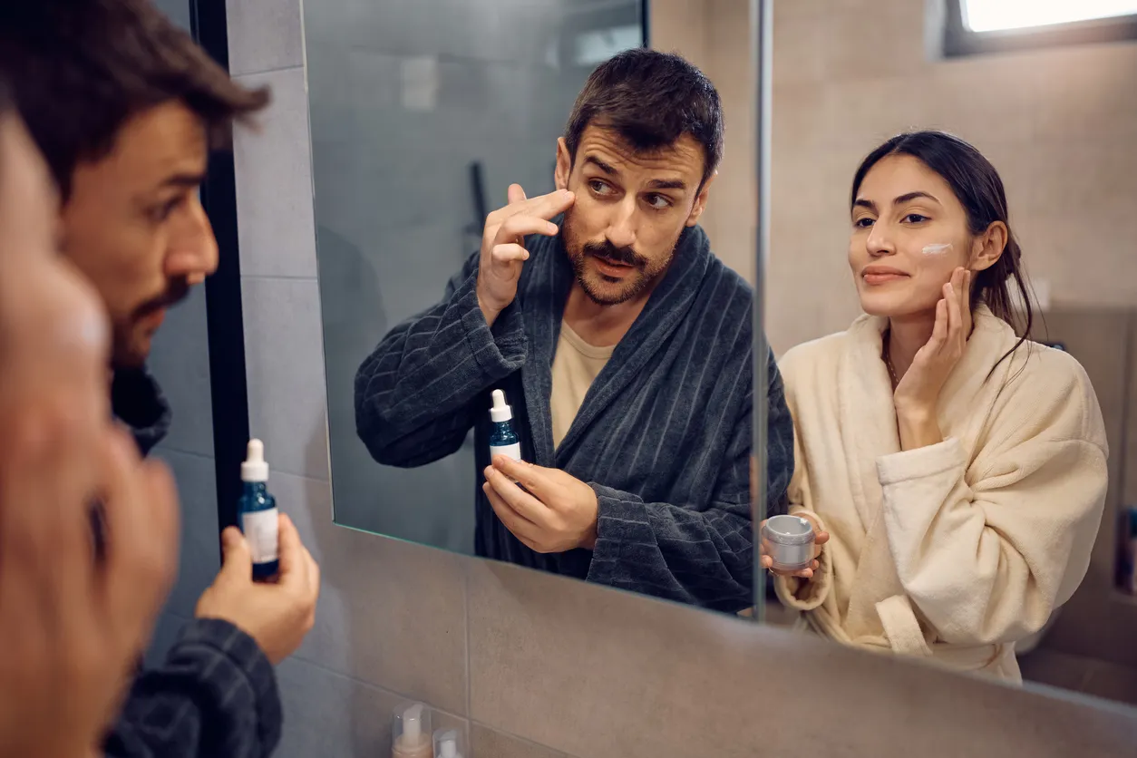 Çift aynada serum ve krem uygularken cilt bakım rutini yapan kadın ve erkek, akne ve gözenek görünümüne yönelik aktif içerikler kullanıyor.