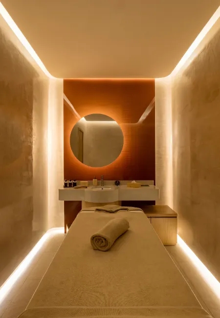 İstanbul'daki en iyi SPA merkezlerinden birinde, vücudu ve zihni dinlendirmek için hazır bekleyen, sıcak renklerle aydınlatılmış modern bir terapi odasında, masaj yatağının üzerinde rulo yapılmış havlu ve arka planda aynalı lavabo ünitesiyle huzurlu bir atmosfer sunuluyor.