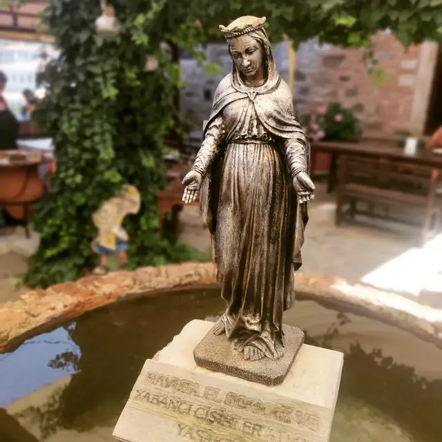 Şirince'deki tarihi St. John the Baptist Kilisesi çevresindeki atmosferi yansıtan, tacıyla ve açık avuçlarıyla duran, bronz görünümlü bir Meryem Ana heykeli, üzerinde havuza el sokmayı ve yabancı cisimleri yasaklayan bir uyarının bulunduğu bir kaide üzerinde açık hava havuzunun ortasında yer alıyor. Arka planda yemyeşil sarmaşıklar ve taş yapılarla bezeli, hafif bulanık bir dış mekan kafe ortamı görünüyor.