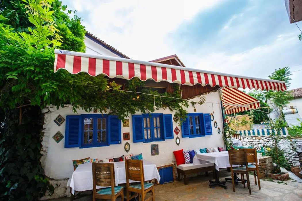 Şirince'nin kendine özgü atmosferini yansıtan bu butik otelin bembeyaz dış duvarları, mavi panjurları ve kırmızı beyaz çizgili tenteleri altında, rengarenk yastıklarla süslenmiş masa ve sandalyelerin bulunduğu davetkar bir açık hava oturma alanı görülüyor.