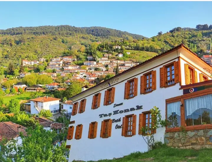 Ön planda, yeşil bir yamacın üzerinde yer alan, ahşap panjurlu pencereleri ve Şirince Taş Konak Butik Otel yazısı bulunan beyaz renkli geleneksel bir yapı görülürken, arka planda yemyeşil ağaçlarla kaplı tepelere yayılmış çok sayıda şirin köy evi ve berrak mavi gökyüzü dikkat çekmektedir.