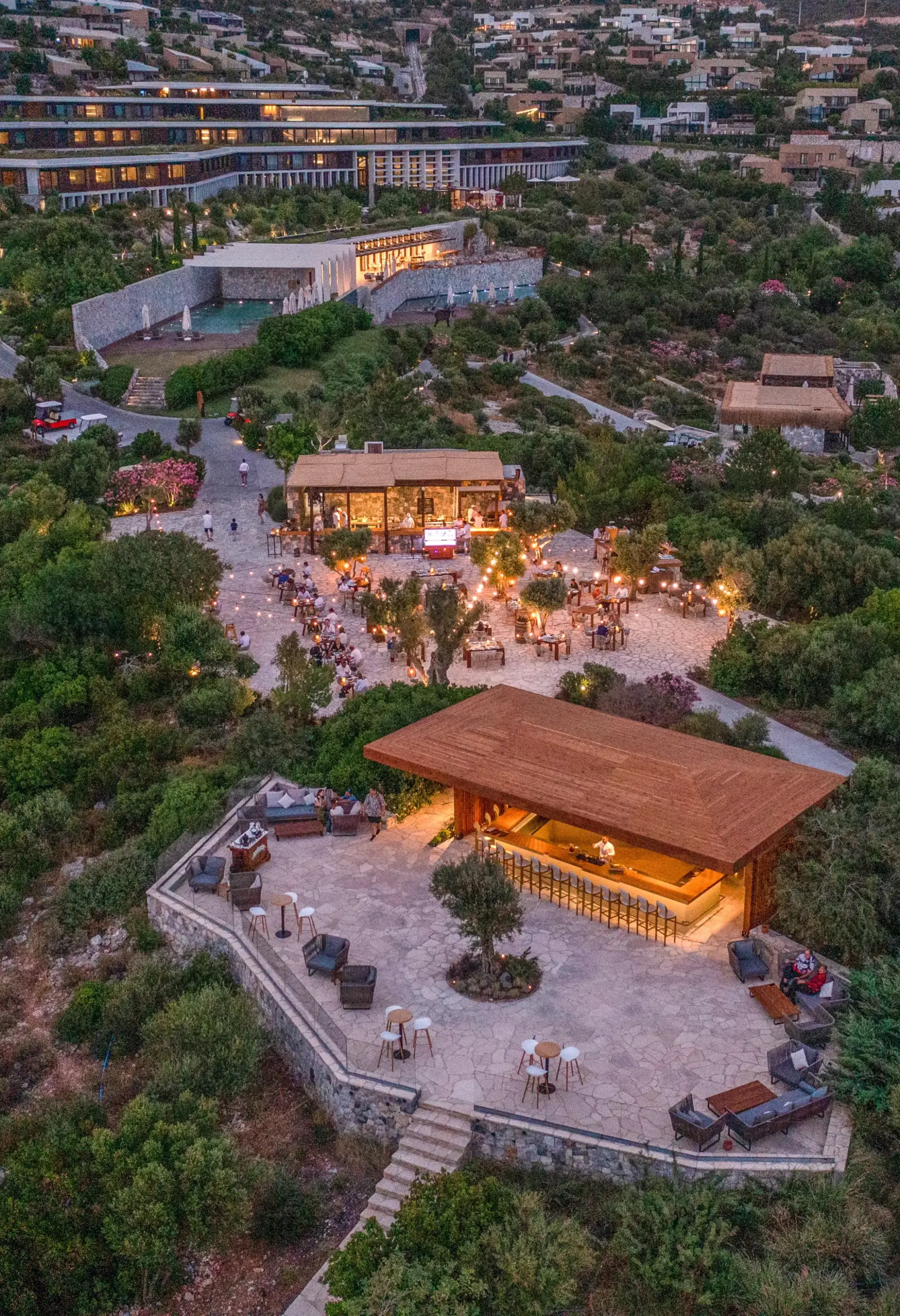 Gün batımında havadan çekilmiş lüks resort alanı; taş teraslı açık hava barı ve ışıklarla aydınlatılmış restoran bölümü, çevresinde zeytin ağaçları ve yamaç boyunca konumlanan modern otel yapılarıyla birlikte.