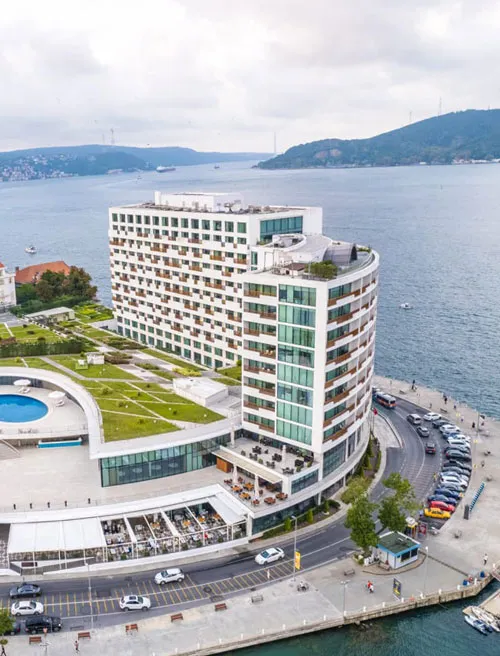 Gri bir gökyüzünün altında, Tarabya sahilinde yer alan modern, çok katlı beyaz otel kompleksi, yeşil teraslı bahçeleri ve açık havuzu ile öne çıkıyor; önünde yoğun bir yol ve park etmiş araçlar, uzakta ise tüm ihtişamıyla Boğaz manzarası görünmektedir.