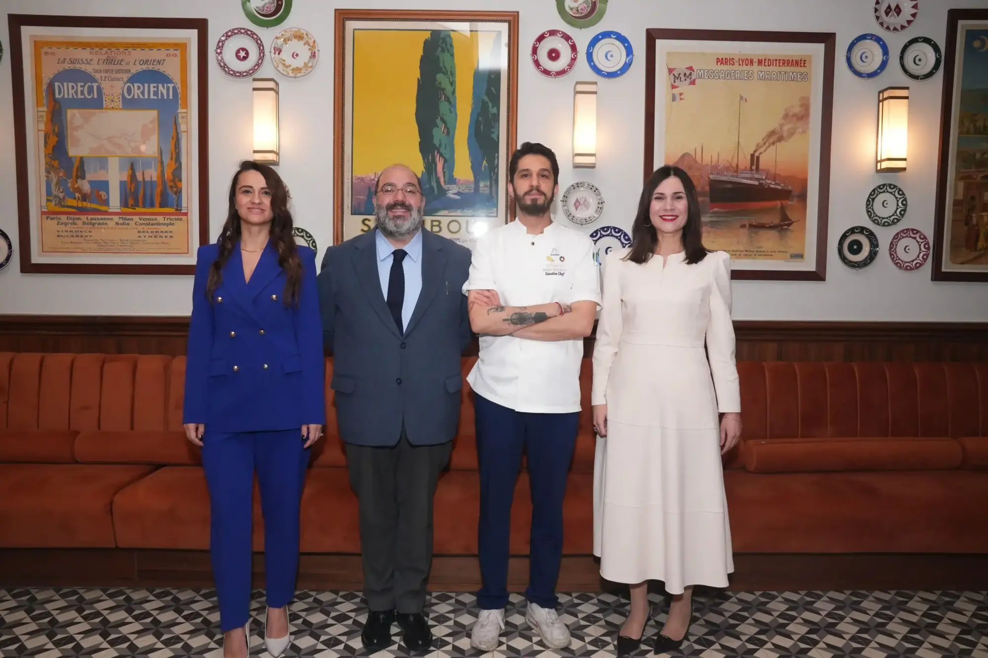Dört kişi yan yana poz veriyor; solda mavi takım elbiseli bir kadın, yanında lacivert takım elbiseli ve kravatlı bir erkek, üçüncü sırada beyaz şef ceketi giymiş bir erkek ve en sağda krem rengi uzun elbiseli bir kadın. Arka planda vintage seyahat afişleri, dekoratif tabaklar ve duvar aplikleri bulunan şık bir restoran ortamı, önlerinde turuncu kadife koltuk yer alıyor.