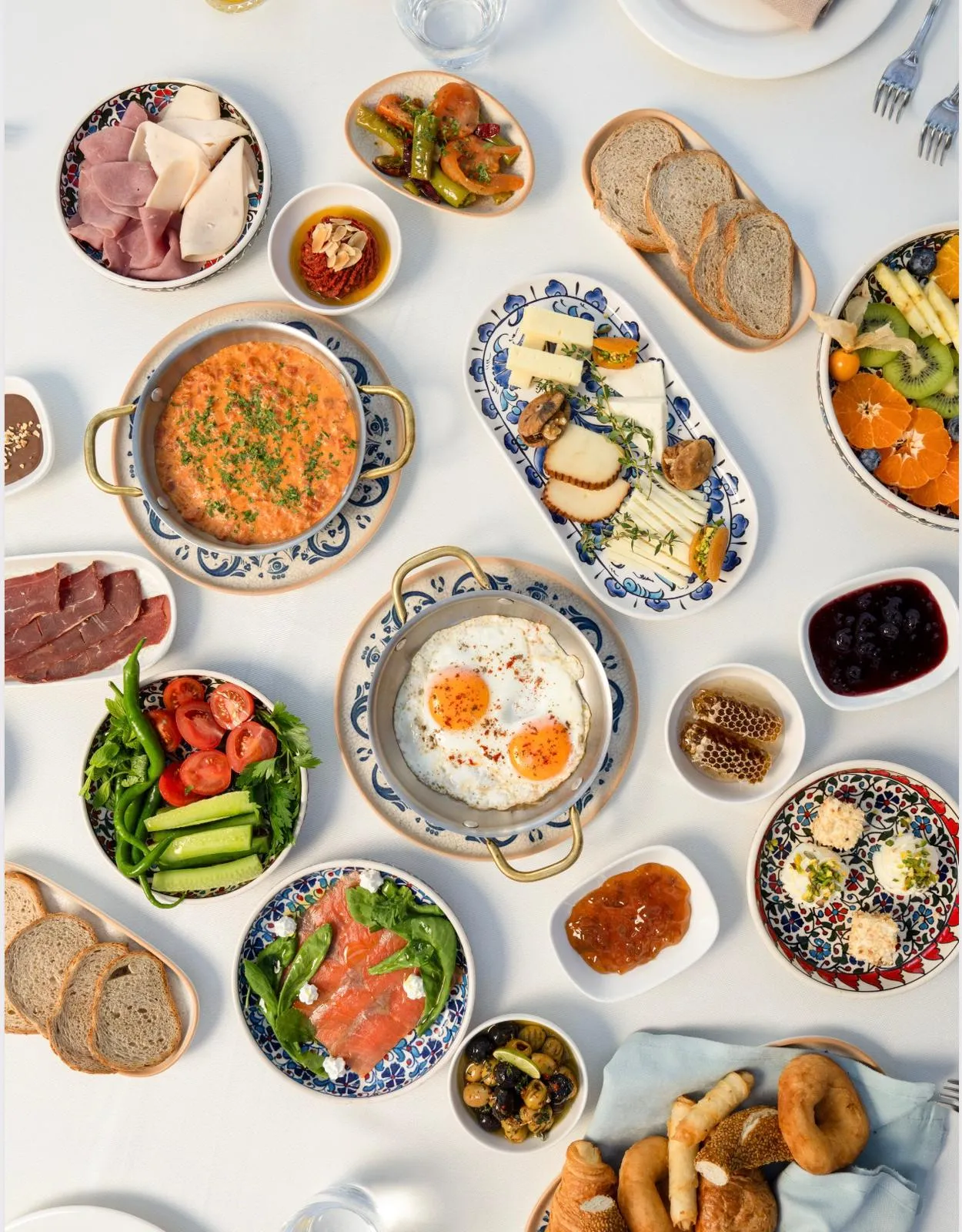 Beyaz bir masa üzerinde, menemen, sahanda yumurta, çeşitli yöresel peynirler, şarküteri ürünleri, taze yeşillik ve domates salatası, zeytin, bal peteği, farklı reçeller, taze ekmekler, meyve tabağı ve simit gibi birçok geleneksel Türk kahvaltısı lezzetini bir araya getiren zengin ve iştah açıcı bir serpme kahvaltı sofrası.
