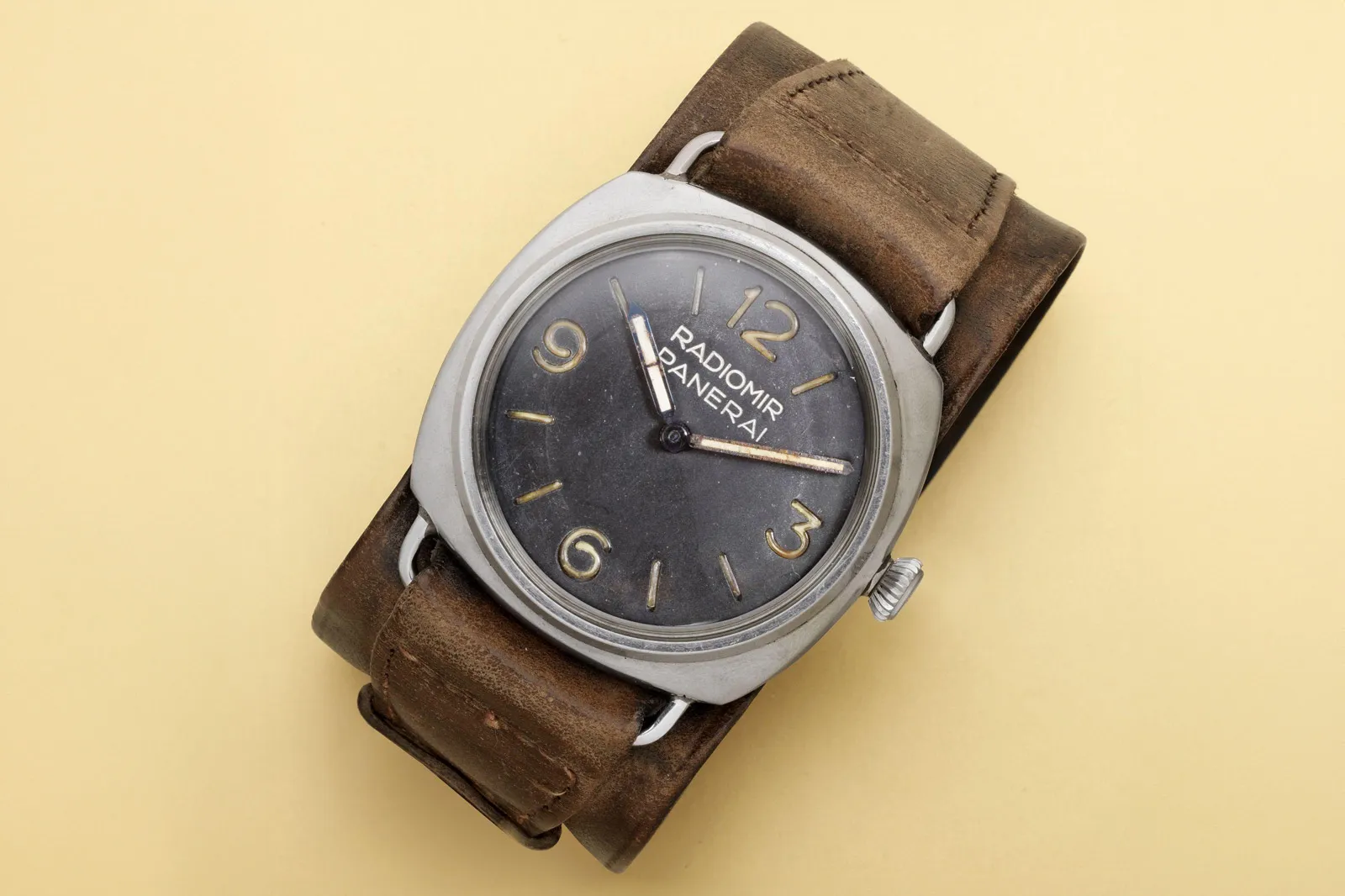 Açık sarı zeminde duran, 1940'lardan kalma, Rolex tarafından Panerai için üretilmiş Panerai Radiomir 3646 model vintage bir kol saati; geniş kahverengi deri kayışıyla, yıpranmış gümüş rengi kasasıyla, karanlık kadranındaki soluk sarı Radiomir Panerai yazısı ve büyük rakamlarıyla öne çıkıyor.