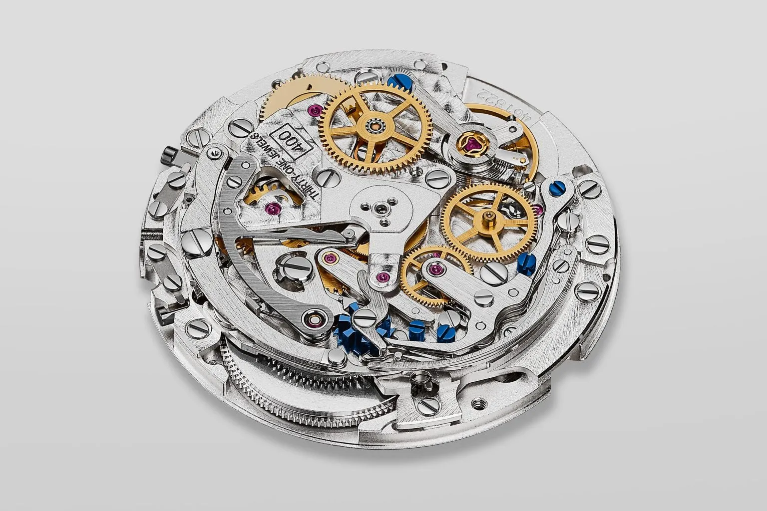 Detaylı bir şekilde yakın çekimle gösterilen Zenith El Primero kronograf mekanizması, karmaşık yapısını oluşturan iç içe geçmiş gümüş rengi köprüleri, parlak altın rengi dişlileri, hassas kırmızı yakut taşları ve bazı parlak mavi vidaları gözler önüne sererek yüksek saatçilik sanatını yansıtıyor.