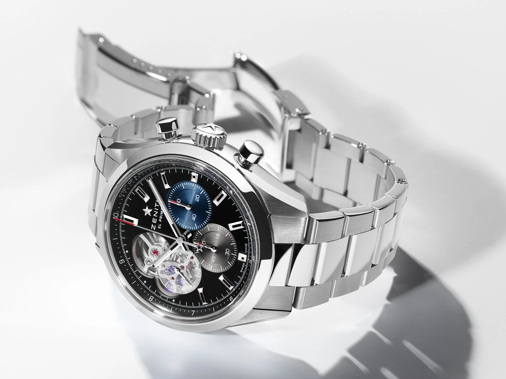 Koyu renk kadranında belirgin mavi ve gri kronograf alt kadranları ile eşsiz açık kalp tasarımı sayesinde mekanik iskeletini sergileyen Zenith Chronomaster Open lüks kol saati, gümüş rengi metal bileziğiyle beyaz zeminde dikkat çekiyor.