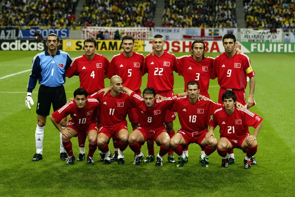 2002 Dünya Kupası'nda yarışan Türkiye milli futbol takımı oyuncuları, bir kaleci mavi formasıyla diğerleri kırmızı formalarıyla olmak üzere yeşil sahada iki sıra halinde poz veriyor, arka planda tribünler ve reklam panoları net bir şekilde seçiliyor.