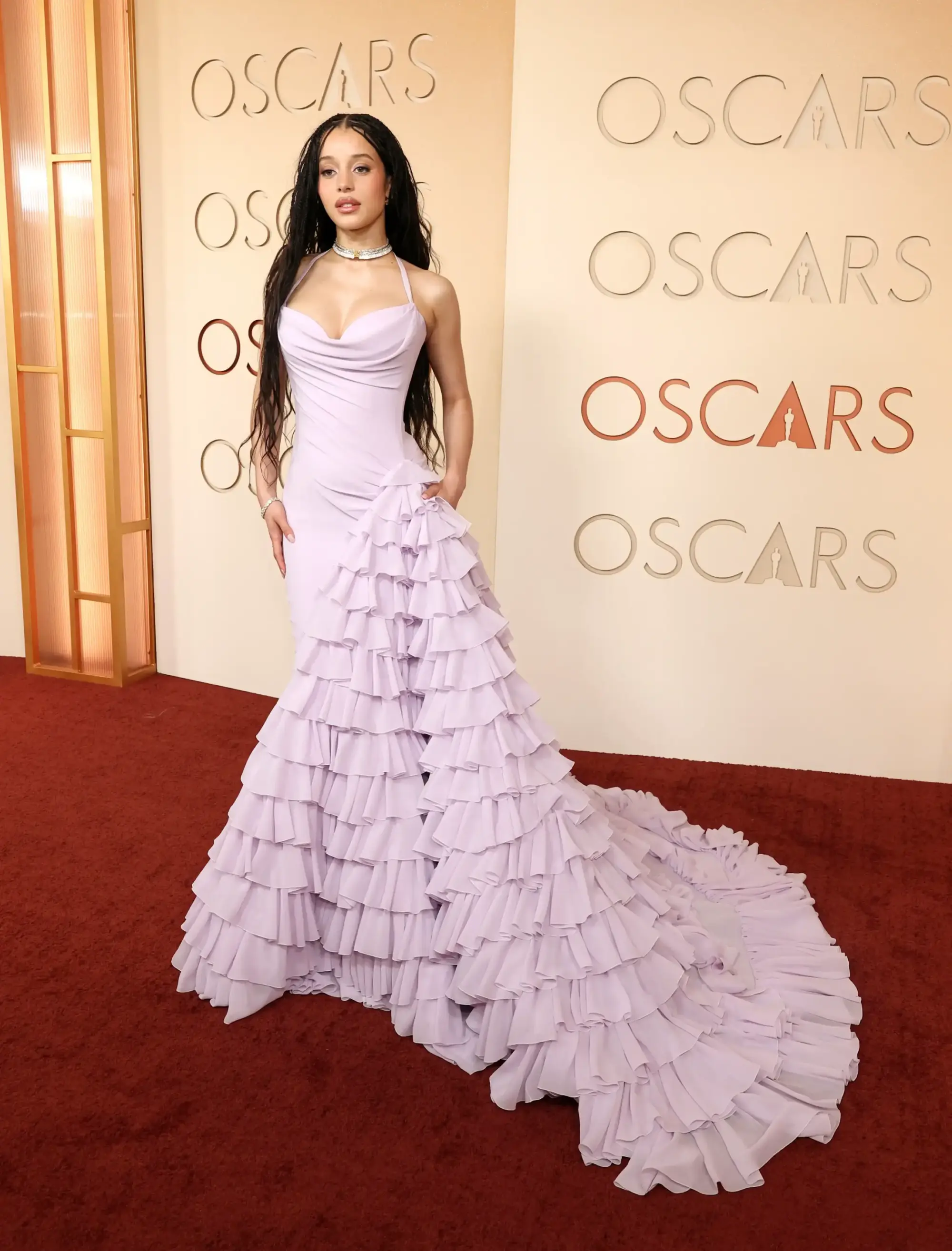 Chase Infiniti, 2026 Oscar kırmızı halısında, ten rengi zeminde OSCARS yazıları olan duvarın önünde dururken, ince askılı, drapeli yaka detaylı, uçuşan kat kat leylak rengi fırfırlarla bezeli uzun couture elbisesi içinde, gösterişli kolyesi ve bileziğiyle tamamladığı şık ve zarif stilini gözler önüne seriyor.