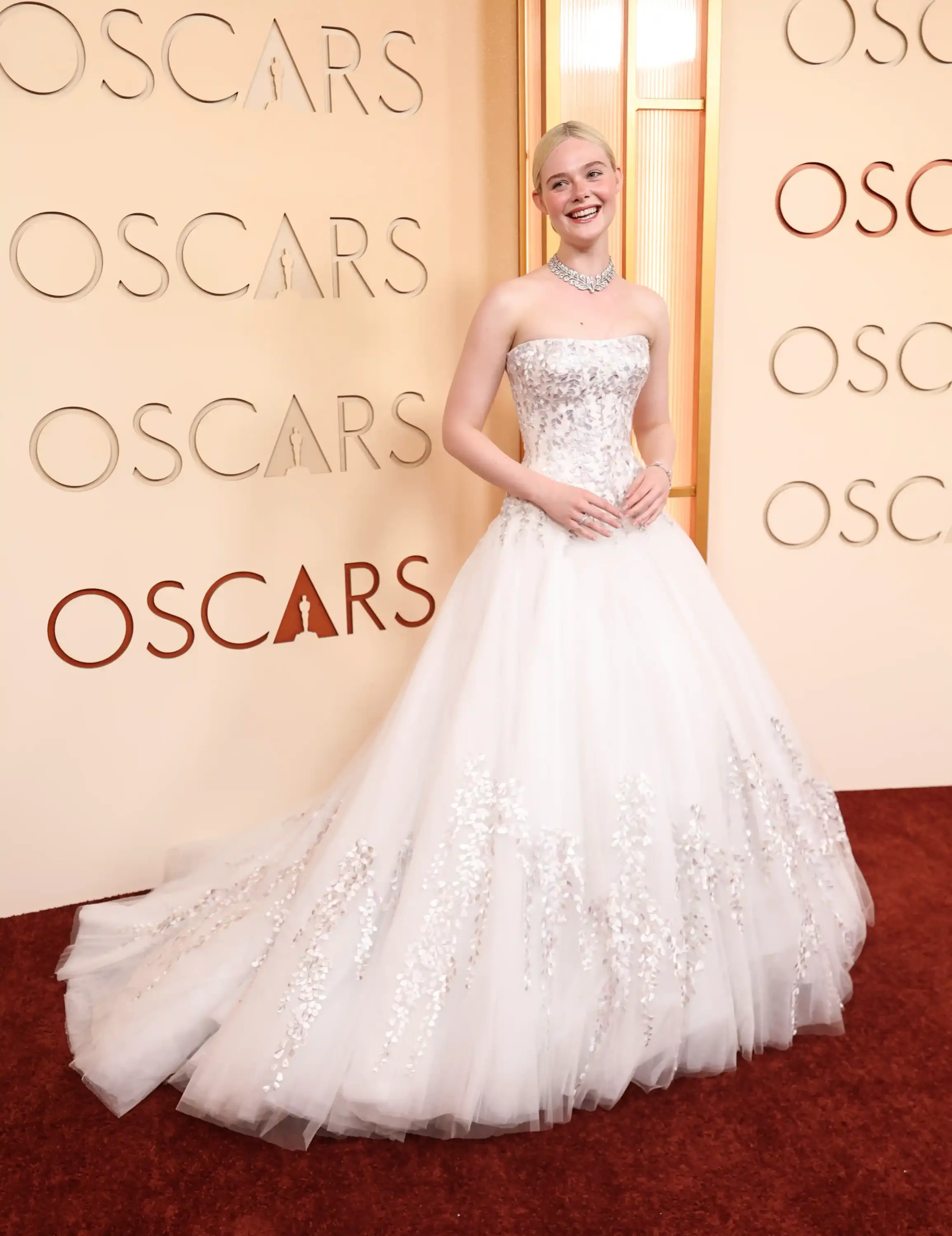 Elle Fanning, 2026 Oscar kırmızı halısı gecesinde, gümüş yaprak motifleriyle işlenmiş straplez, kabarık beyaz couture elbisesi ve parlak pırlanta kolyesiyle şık bir görünüm sergileyerek, Oscar logolu duvar önünde gülümsüyor.