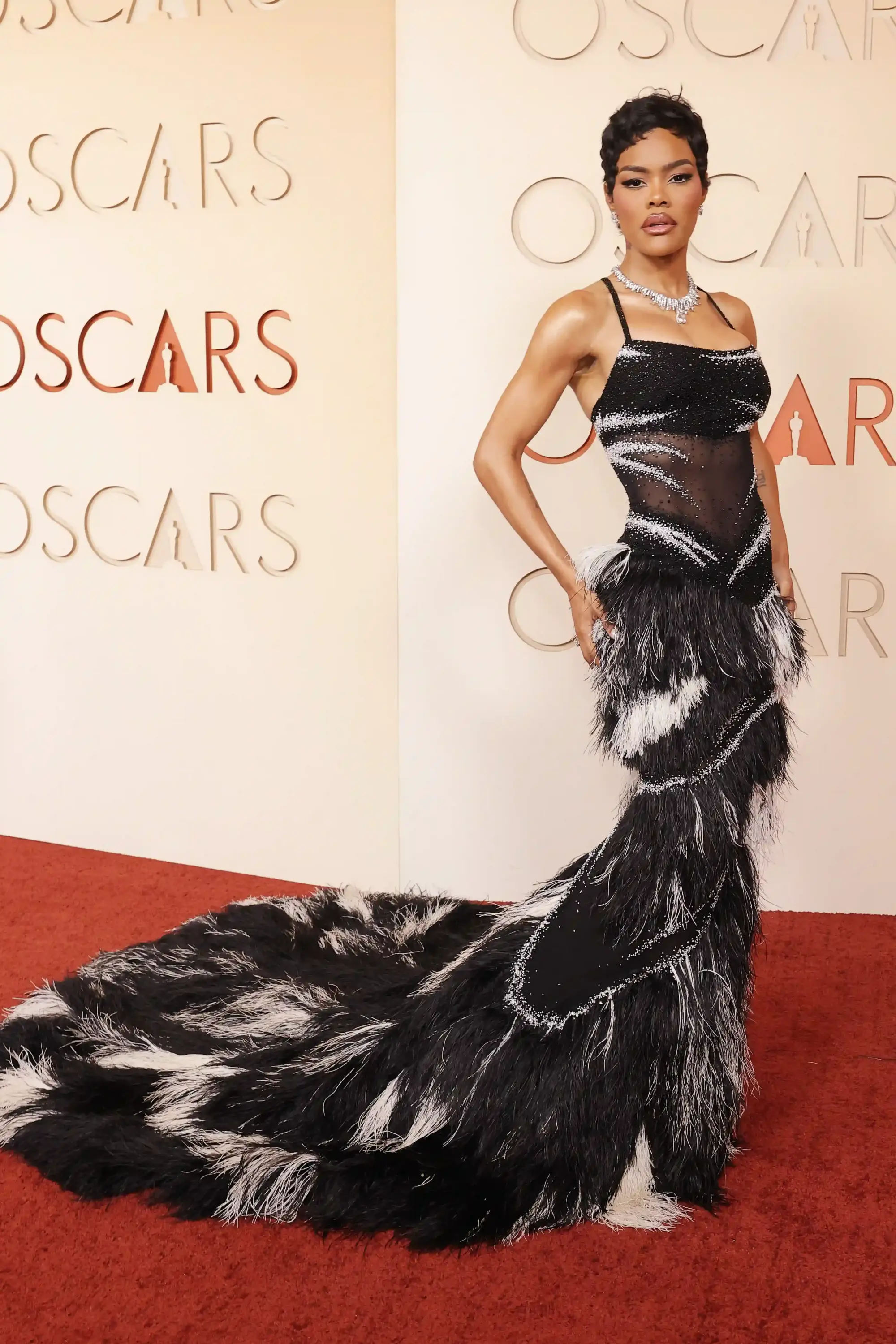 Teyana Taylor, 2026 Oscar kırmızı halısında, siyah ve beyaz tüylerle bezeli, transparan işlemeli ve uzun kuyruklu couture tasarımı elbisesiyle, pırlanta mücevher seçimleriyle stilini ortaya koyarak öne çıkan görünümler arasında yer alıyor.