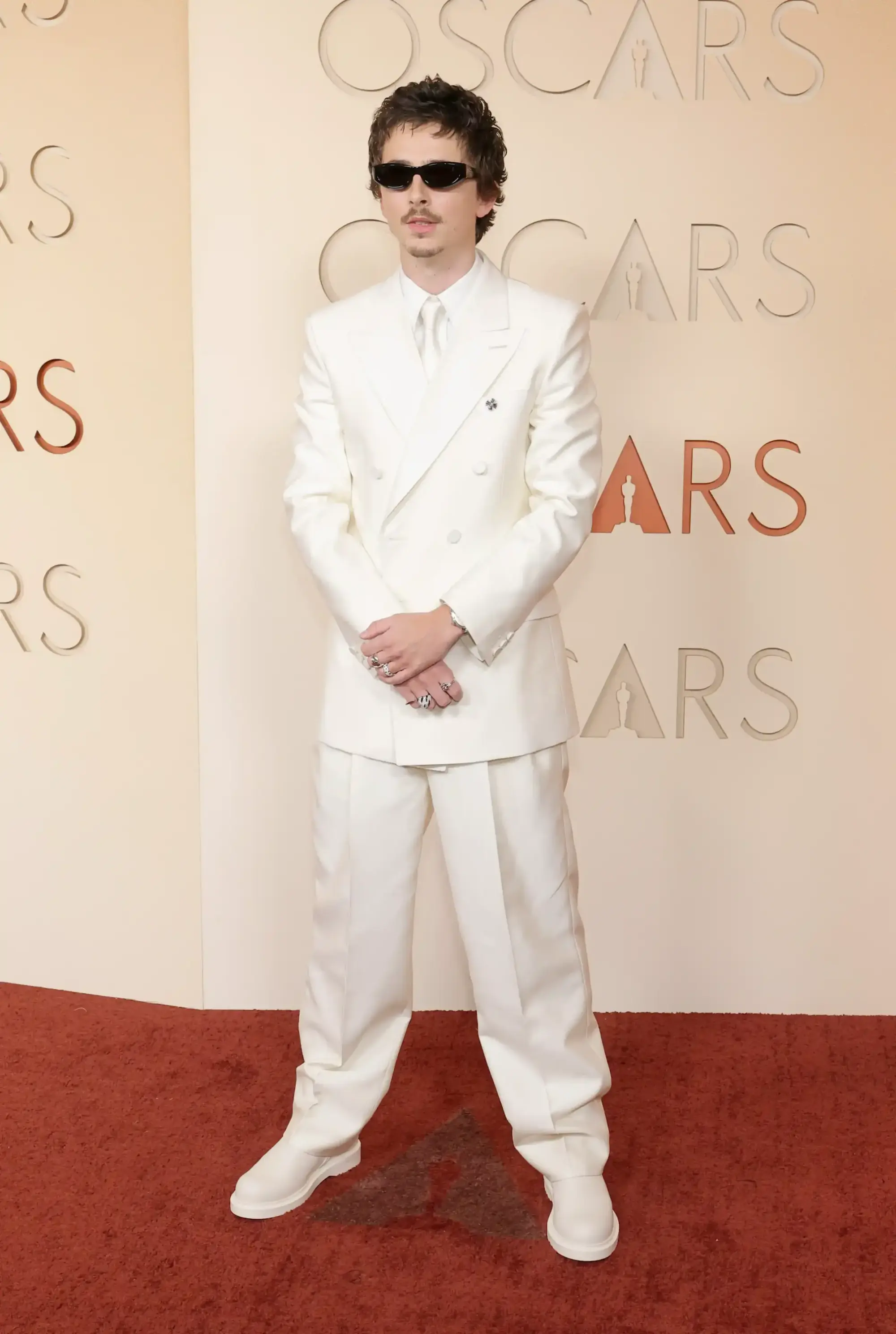 Timothée Chalamet, 2026 Oscar kırmızı halısında, ten rengi zemin üzerinde kabartma Oscar logosunun bulunduğu bir duvarda poz verirken, uyumlu beyaz renkteki çift düğmeli takımı, kalın tabanlı botları, siyah dar çerçeveli güneş gözlükleri ve yüzükleriyle gecenin öne çıkan şık görünümlerinden birini sergiliyor.