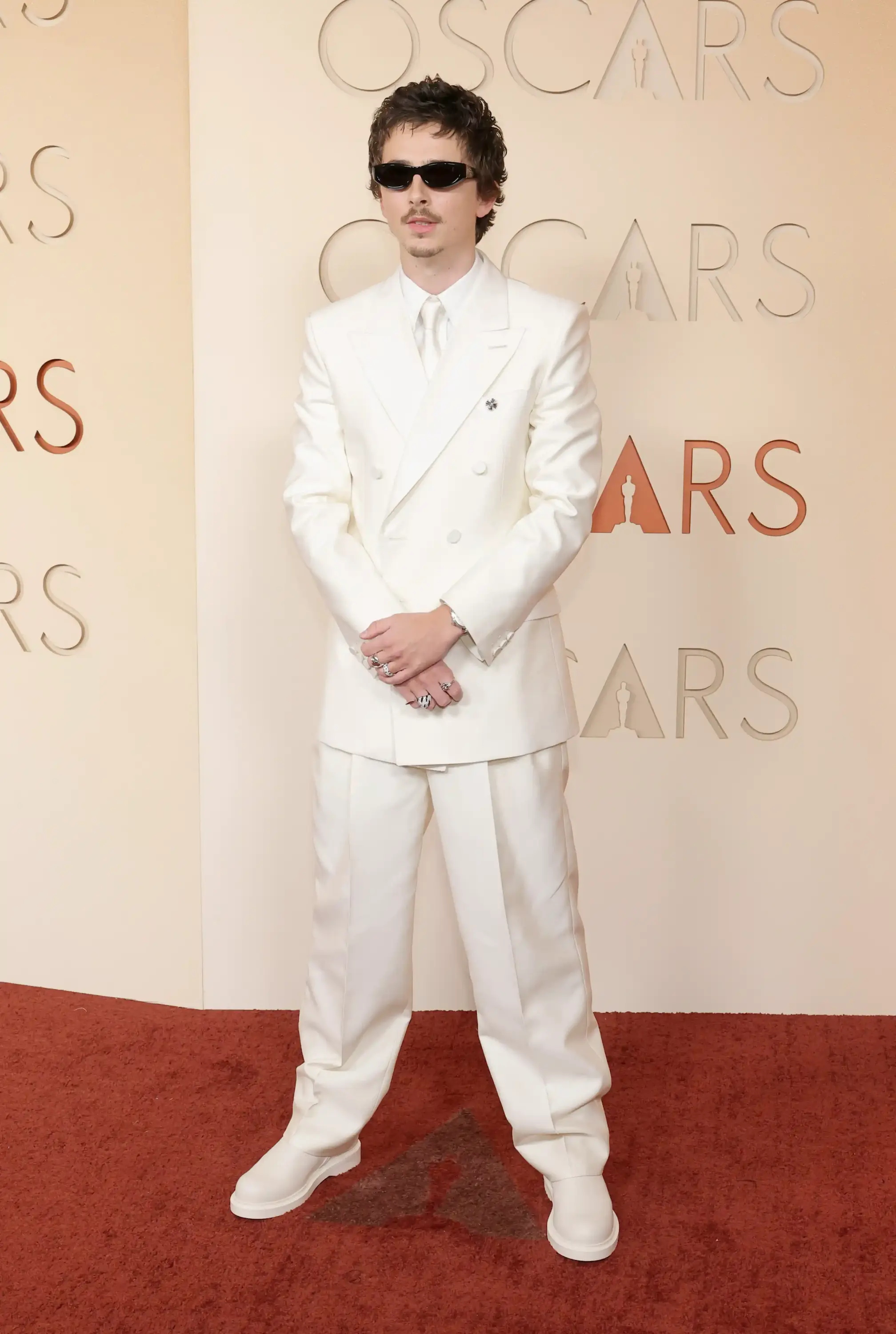 Timothée Chalamet, 2026 Oscar kırmızı halısında, ten rengi zemin üzerinde kabartma Oscar logosunun bulunduğu bir duvarda poz verirken, uyumlu beyaz renkteki çift düğmeli takımı, kalın tabanlı botları, siyah dar çerçeveli güneş gözlükleri ve yüzükleriyle gecenin öne çıkan şık görünümlerinden birini sergiliyor.