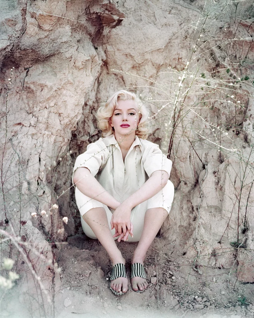 Marilyn Monroe, ikonik 60'lar bob saç modeli ve kırmızı rujuyla, bej renkli gömlek ve pantolon takımı ile çizgili sandaletleriyle kayalık, kurak bir zeminde bağdaş kurmuş oturmuş, kameraya bakıyor.