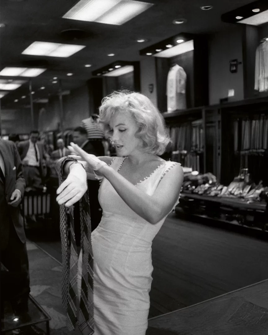 Siyah beyaz karede, 60'lar bob saç modeliyle ikonik sarı saçlı Marilyn Monroe, beyaz eldivenleri ve açık renk elbisesiyle bir erkek giyim mağazasında desenli bir kravatı dikkatle incelerken, arka planda takım elbiseli figürler ve vitrinler seçiliyor.