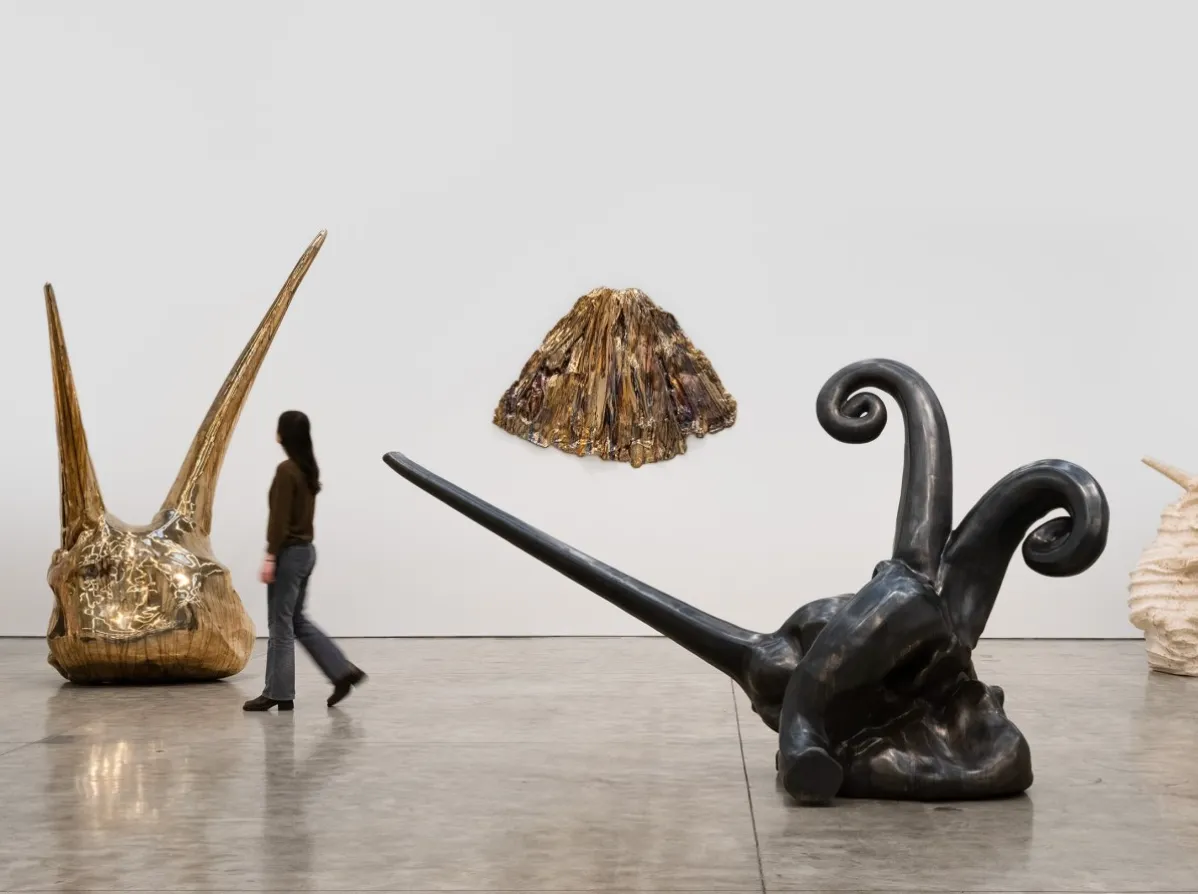 Beyaz duvarlı, çağdaş bir sanat sergisinde, sol tarafta sivri uçlu iki boynuza sahip, altın rengi, dokulu büyük bir heykelin ve sağ tarafta yerde yatan, kıvrımlı kolları olan koyu renkli, organik formlu bir heykelin arasında yürüyen bir kadın görülüyor, arka planda ise duvarda parıldayan konik bir sanat eseri asılı duruyor.
