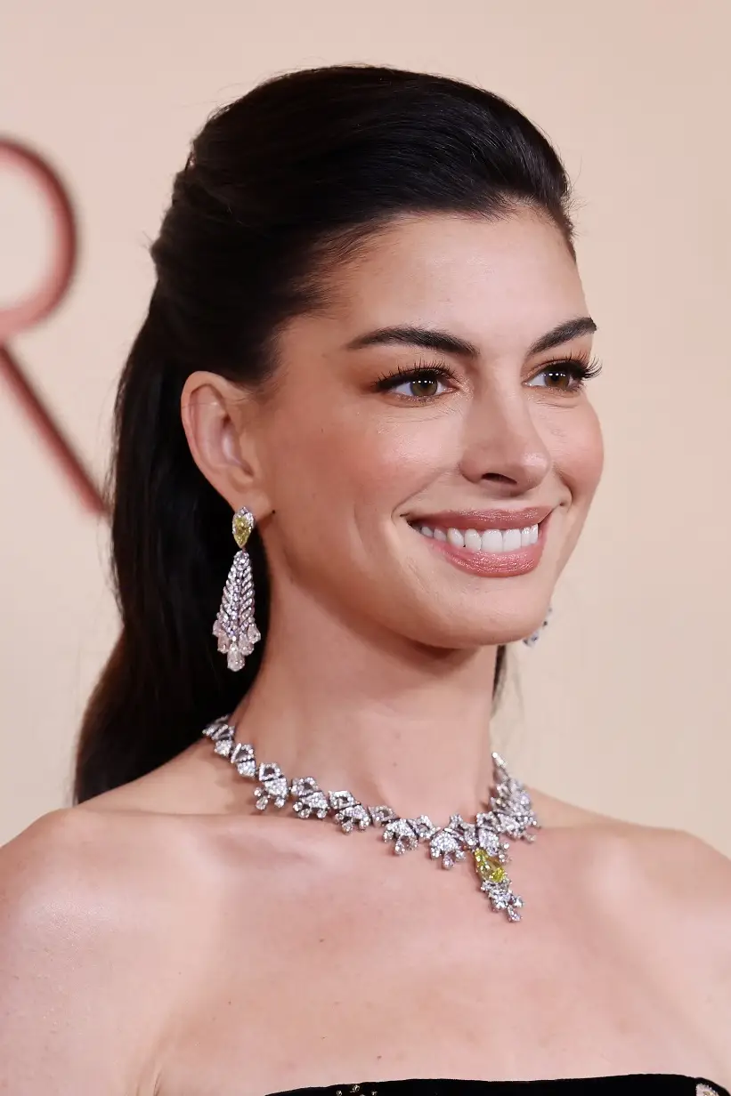 Çarpıcı elmas ve damla sarı taşlarla bezeli gösterişli kolye ve küpeler takmış Anne Hathaway, Oscar Töreni'nde yarı toplanmış koyu saçları ve doğal makyajıyla zarifçe gülümserken, arka planda hafif bulanık bir salon detayı görünüyor.