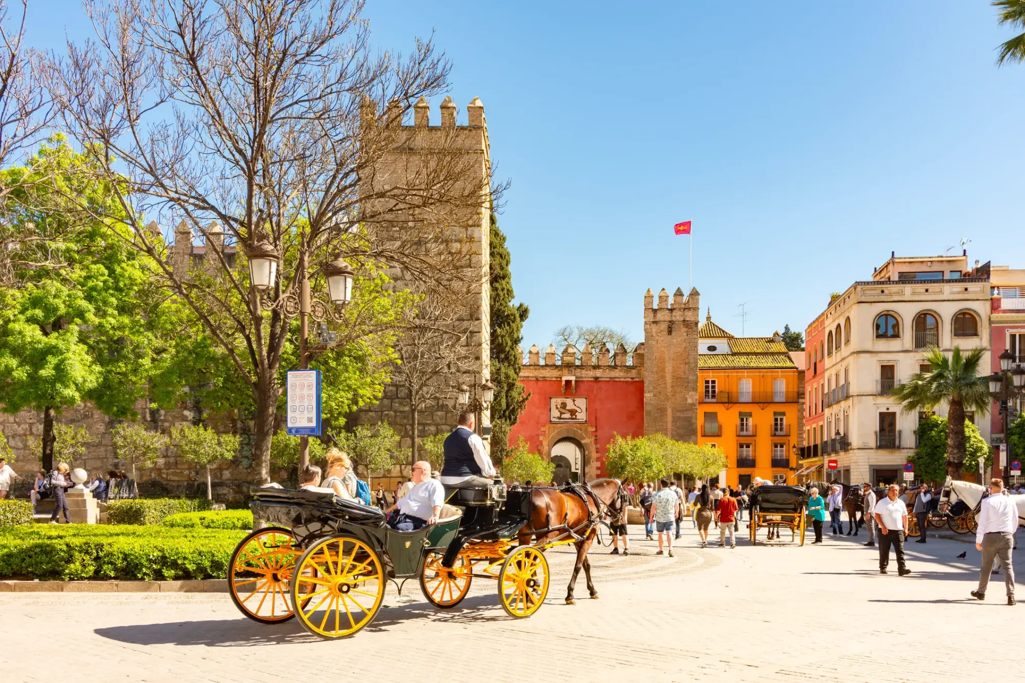 Güneşli bir günde, İspanya'nın tarihi Sevilla şehrinde, Archivo de Indias çevresindeki canlı meydanda, atlı faytonlarla gezintiye çıkan turistler ve yürüyen yayalar arasında hareketli bir yaşam sahnesi resmediliyor; solda yükselen, mazgallı taş duvarlara ve aslan armalı kemerli kırmızı bir kapıya sahip tarihi bir yapı dikkat çekiyor.