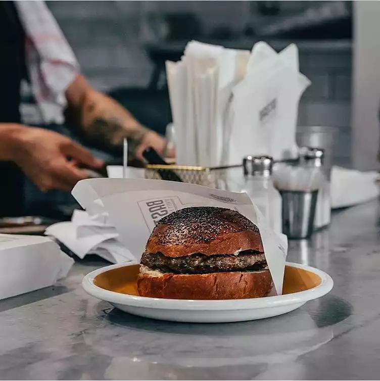 Şefin favorilerinden Basta Moda'da mermer tezgah üzerinde beyaz tabakta sunulan, özel kağıdına sarılı, haşhaşlı parlak ekmekli ve kalın köfteli sıra dışı hamburger. Arka planda dövmeli kollarla yemek hazırlayan bir aşçının silüeti