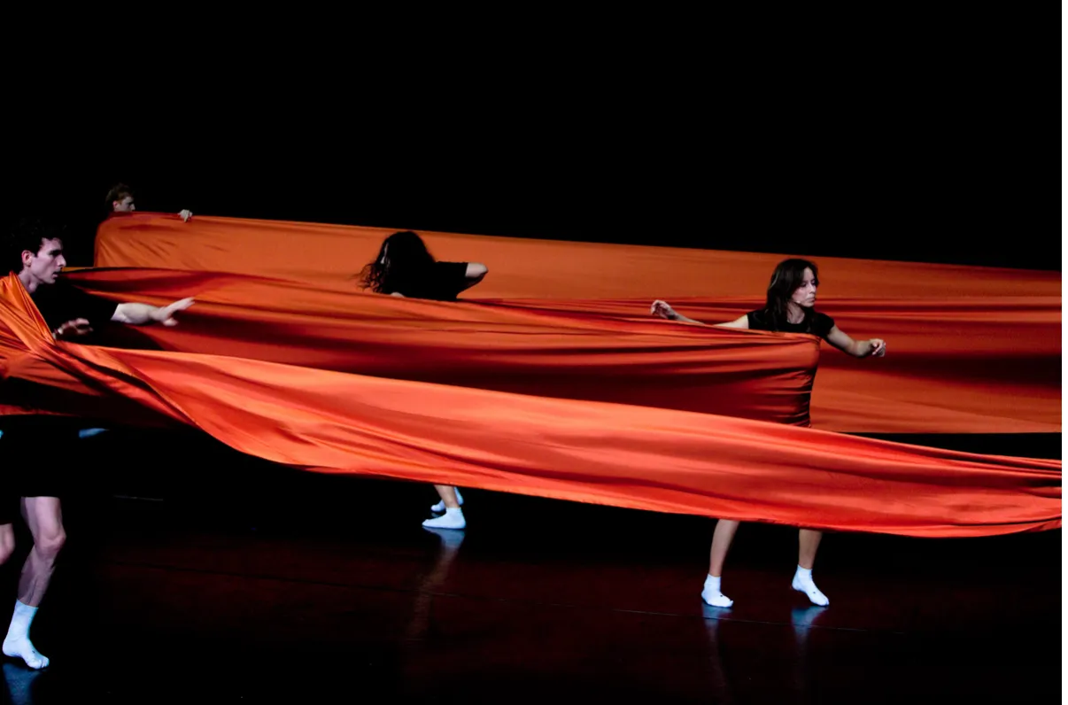 Karanlık bir sahnede, birkaç performansçı büyük, akıcı turuncu bir kumaşı dalgalandırarak ve vücutlarının etrafında şekillendirerek dinamik bir hareket sergiliyor; bu etkileyici görsel, Belçika Pavyonu'nda sunulan yenilikçi bir gösteriden bir anı yansıtıyor.