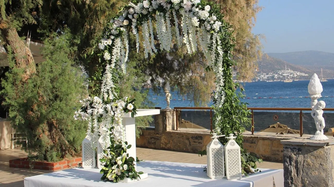 Bodrum'un masmavi denizi ve yemyeşil doğasıyla iç içe, beyaz çiçekler ve yeşil yapraklarla süslenmiş şık bir düğün kemeri, uzakta teknelerin göründüğü bir koy manzarası eşliğinde unutulmaz bir nikah töreni için hazır edilmiş sade ve zarif bir ortam sunuyor.