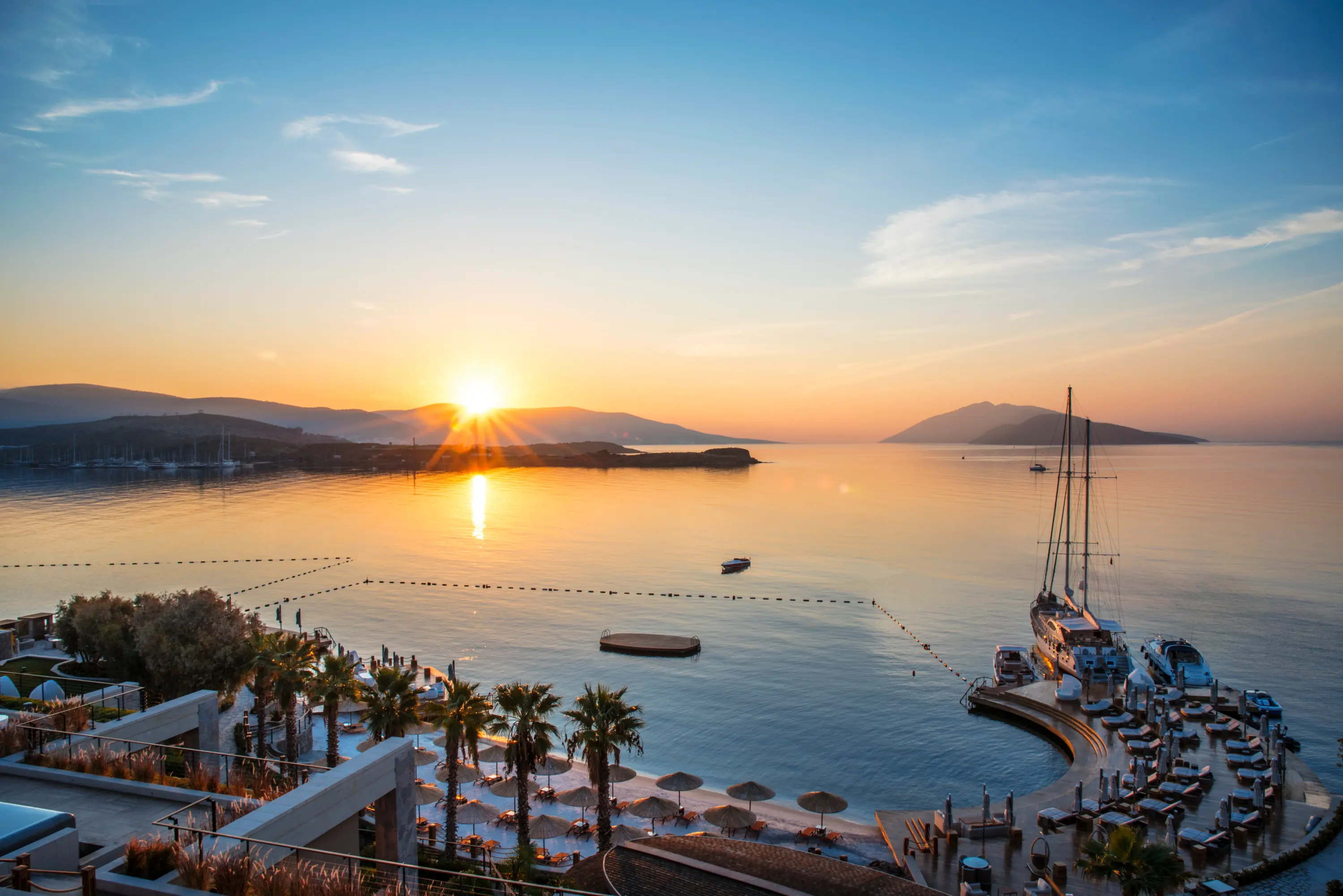 Bodrum'un lüks bir düğün mekanında gün batımının denizin üzerinde oluşturduğu büyüleyici yansımalarla parlayan, palmiye ağaçları ve sahildeki şezlonglar eşliğinde teknelerin demirlediği huzurlu bir atmosfer.