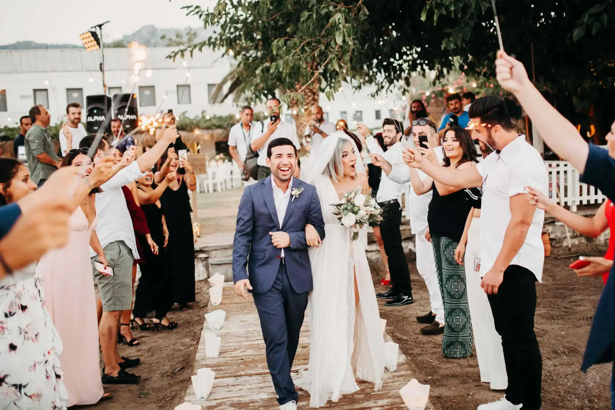 Bodrum'un doğal güzellikleri içinde gerçekleşen unutulmaz bir düğünde, gelin ve damat, yanan maytaplar ve sevgi dolu misafirlerin alkışları eşliğinde, beyaz kağıt fenerlerle aydınlatılmış toprak bir yolda neşe içinde ilerleyerek Bodrum düğün mekânlarının samimi ve kutlamaya değer atmosferini yansıtıyor.