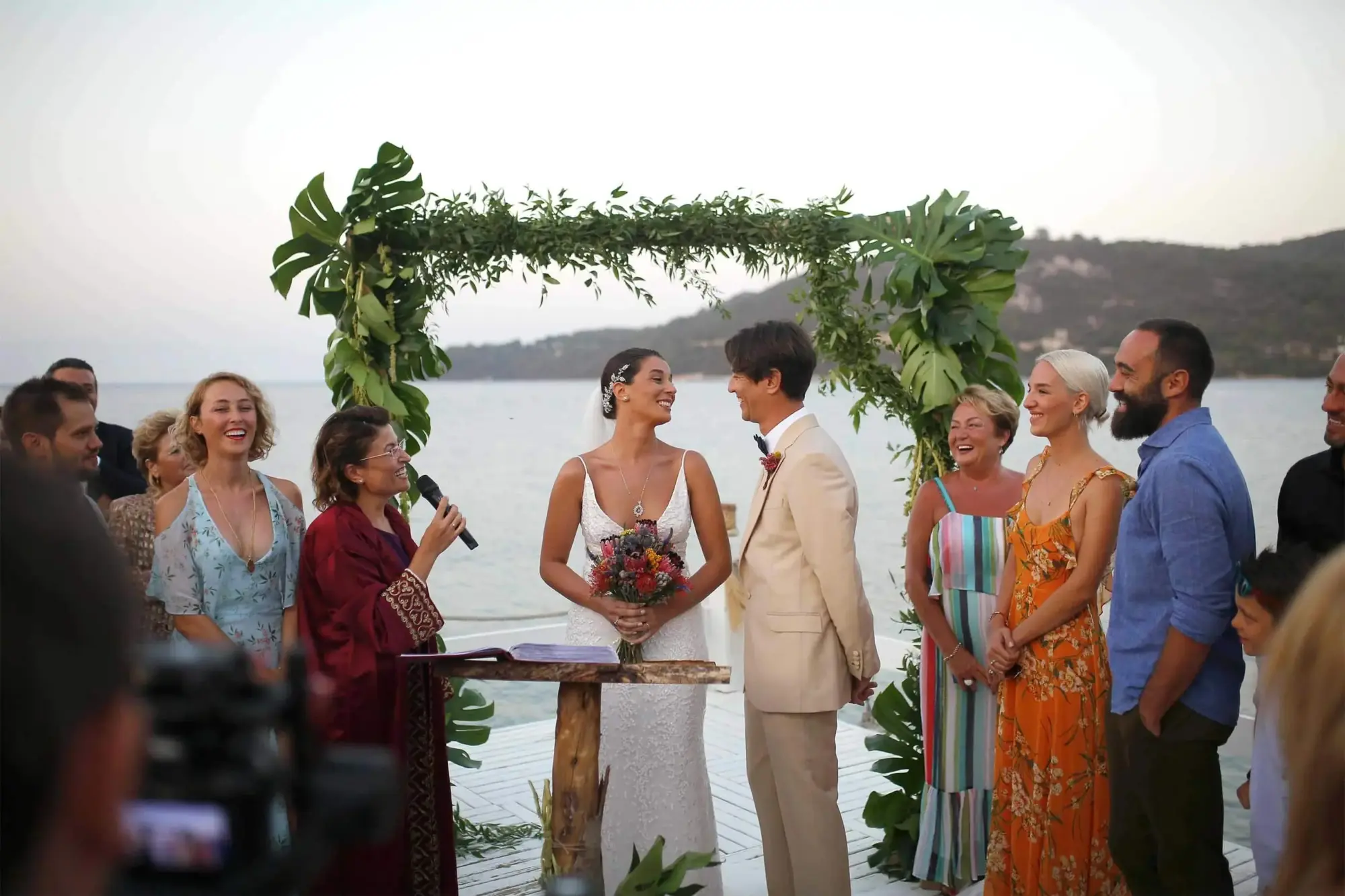 Bodrum'un eşsiz deniz manzarasına karşı, yeşil yapraklı bir takın altında nikah masasında duran gülen gelin ve damat ile onlara mutlulukla bakan konukların olduğu, özel bir düğün töreninin neşeli anı.