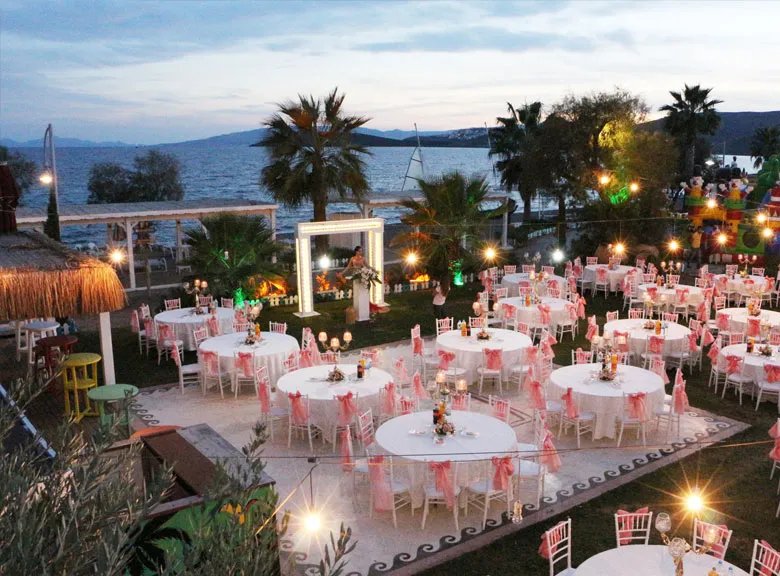 Açık havada, Bodrum'un eşsiz deniz manzarasına sahip düğün mekânlarından birinde, beyaz örtülü ve pembe tüllerle süslü yuvarlak masaların yeşil bir alana özenle dizildiği, hafifçe aydınlatılmış romantik bir akşam kutlaması ortamı gözüküyor.