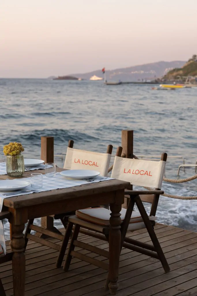Bodrum'da samimi bir düğün mekânı olan La Local'da, denize sıfır ahşap iskelede hazırlanan masada, dalgaların sesi eşliğinde kurulmuş tabaklar, bardaklar ve La Local logolu sandalyelerle romantik bir akşam yemeği manzarası.