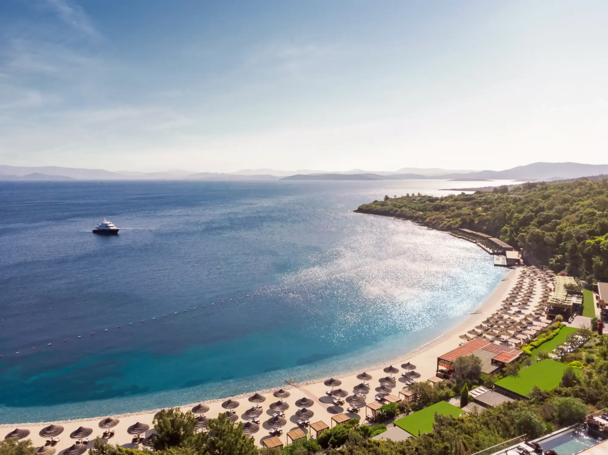 Bodrum'un göz kamaştıran Ege Denizi kıyısında, zarif bir koyda uzanan bej renkli plajında düzenli sıralanmış hasır şemsiyeler ve şezlonglarla donatılmış lüks bir tesisin yemyeşil bahçeleri, uzakta seyreden bir yat eşliğinde rüya gibi düğünlere ev sahipliği yapabilecek büyüleyici bir manzara sunuyor.