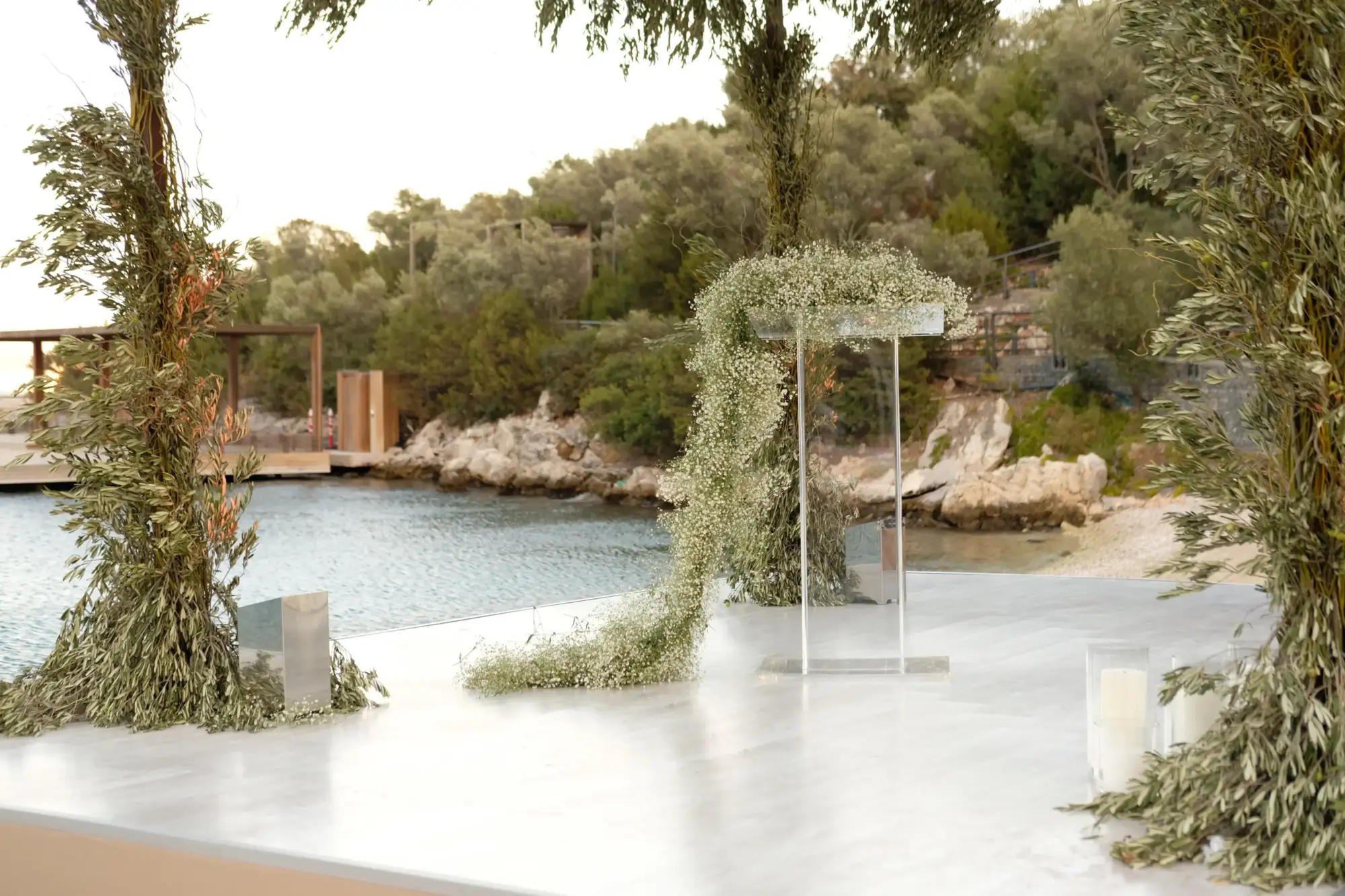 Bodrum'da denize nazır lüks bir düğün mekânının beyaz platformu üzerinde, zeytin dalları ve bembeyaz cipsofillalarla minimalist bir şekilde süslenmiş tören alanı, yeşil tepeler ve kayalık bir koy eşliğinde romantik ve zarif bir atmosfer sunuyor.