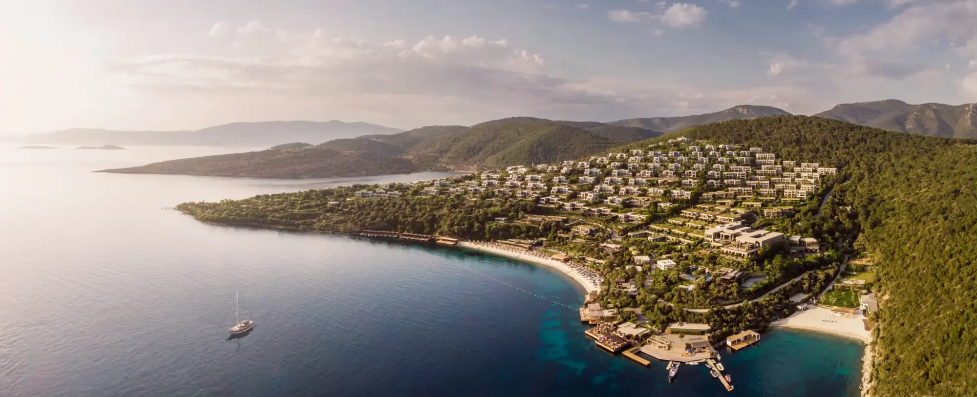 Lüks Mandarin Oriental, Bodrum'un yemyeşil bir yamaçta beyaz villaları, turkuaz renkli denizi, uzun kumlu plajı ve ahşap iskeleleriyle büyüleyici bir Ege koyuna yayıldığı havadan çekilmiş bu geniş görüntü, unutulmaz Bodrum düğün mekanları için eşsiz bir atmosfer sunuyor.