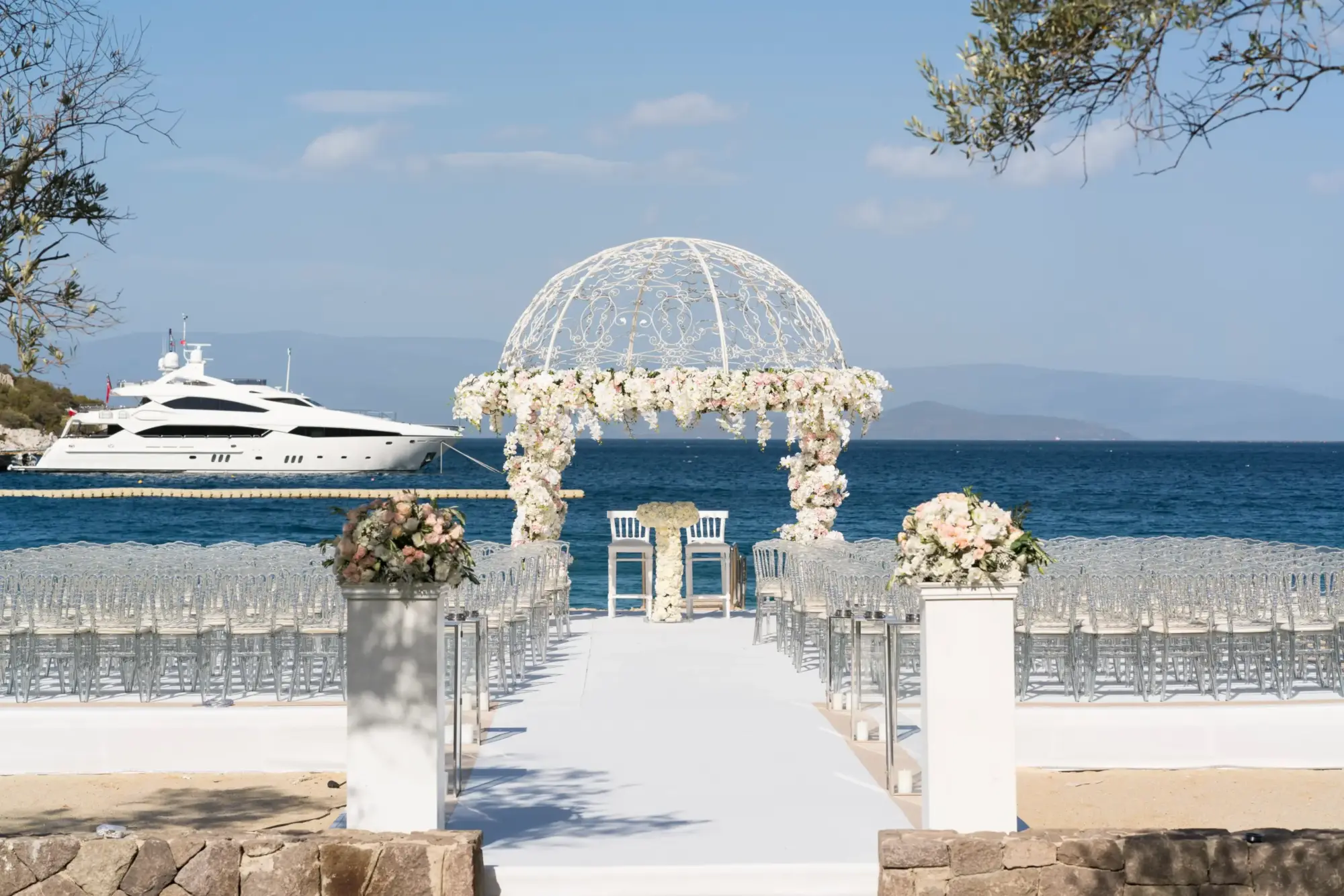 Masmavi Ege Denizi'nin kıyısında, zarif beyaz çiçekler ve hafif pembe güllerle bezenmiş kubbeli bir nikah kemerinin altında, şeffaf sandalyelerin sıralandığı bembeyaz bir yolun denize uzandığı şık bir Bodrum düğün mekanı, lüks bir yatın eşlik ettiği unutulmaz bir tören için hazır.