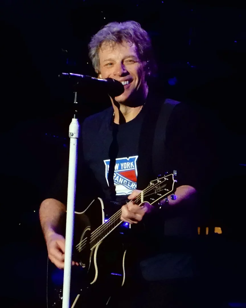 Bon Jovi, Madison Square Garden'da sahne alırken, New York Rangers logolu tişörtüyle gülümseyerek akustik gitarını çalıyor, parlak spot ışıkları altında enerji dolu bir performans sergiliyor.