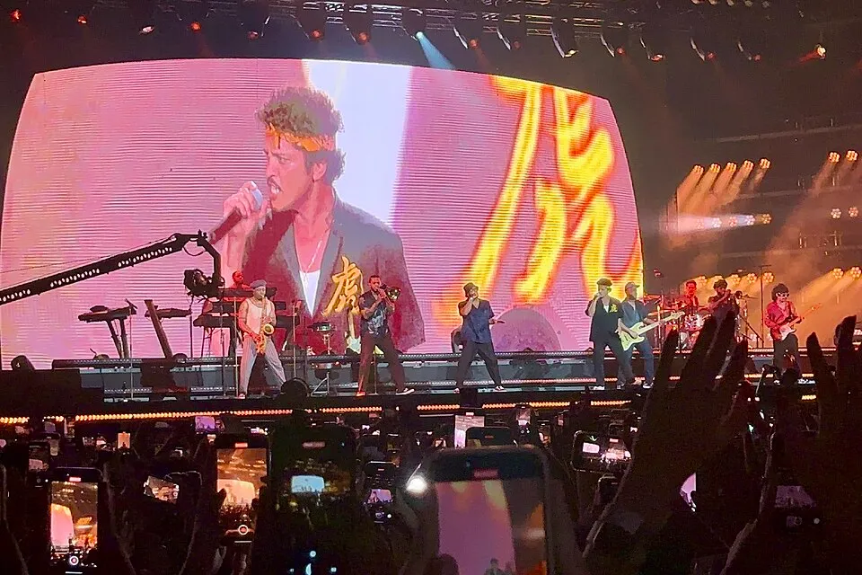 Singapur'daki bir konserde, sahnedeki Bruno Mars dev bir ekranda yakın plandan şarkı söylerken, arkasındaki orkestra çeşitli enstrümanlar çalıyor ve kalabalık telefonlarıyla bu canlı performansı kaydediyor.