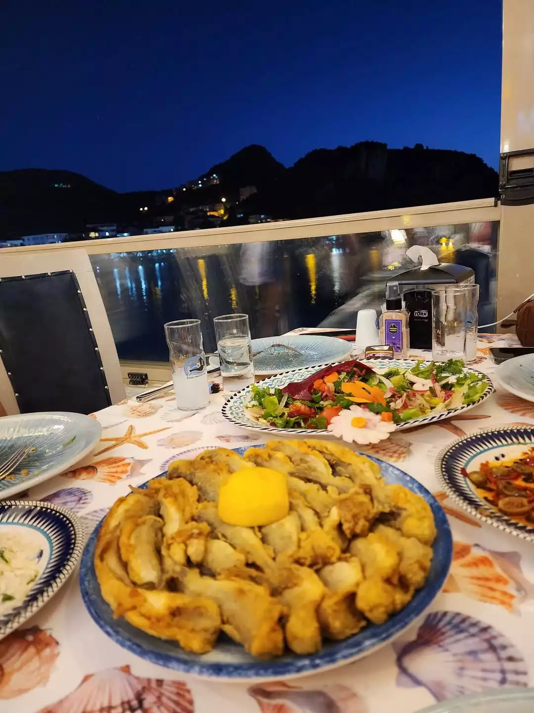 Şefin seyahat etmeye değer bulduğu Bartın Amasra Canlı Balık Restoran'daki deniz manzaralı akşam yemeği. Deniz kabuğu desenli masada taze salata, rakı kadehi ve ortasında limon olan kızarmış balık tabağı gece manzarası eşliğinde