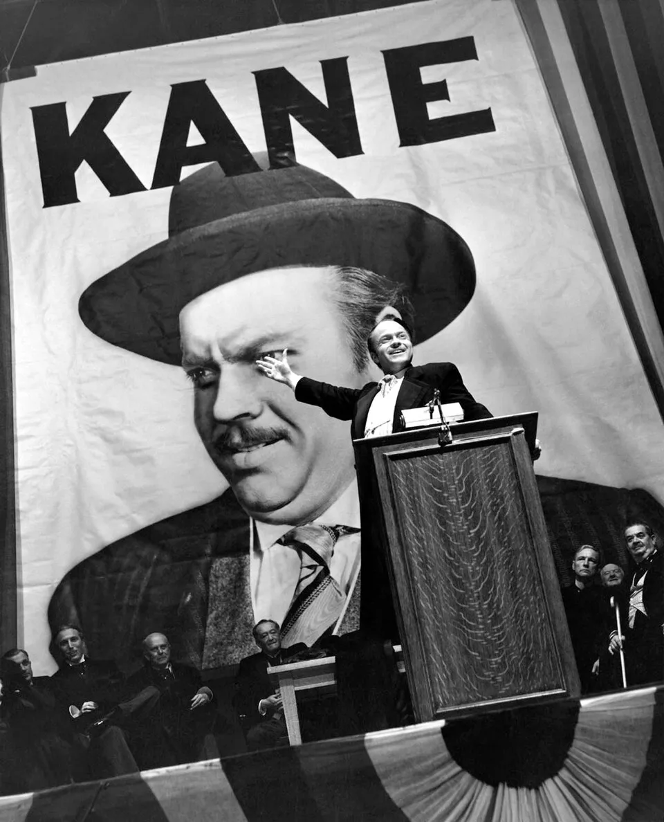 Citizen Kane filminden bir sahnede, Orson Welles'in canlandırdığı Charles Foster Kane, arkasında kendi devasa resminin ve KANE yazısının bulunduğu bir afişin önünde, kürsüde elini havaya kaldırarak neşeyle hitap ederken görülüyor ve bu kare, karakterin politik yükselişini vurguluyor.