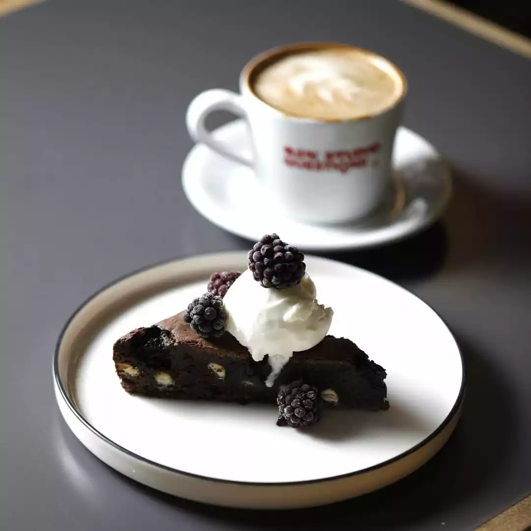 Oggusto, Salvatore Cannetiello – Şeflerin Lezzet Rotaları. Şefin favorisi Coffee Manifesto'yu temsil eden sunum: Gri masa üzerindeki beyaz tabakta, taze böğürtlen ve krema ile taçlandırılmış çikolatalı brownie dilimi öne çıkarken, arka planda latte sanatlı sıcak bir kahve ona eşlik ediyor.