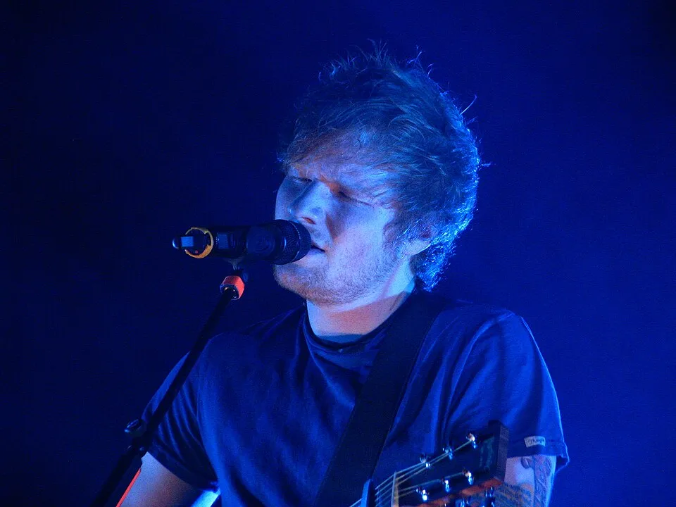 Mavi sahne ışıkları altında parlayan kızıl saçları ve koyu renk tişörtüyle Ed Sheeran, akustik gitarını çalıp mikrofonuna içten bir ifadeyle şarkı söylerken canlı bir konserde derinlemesine bir performans sergiliyor.