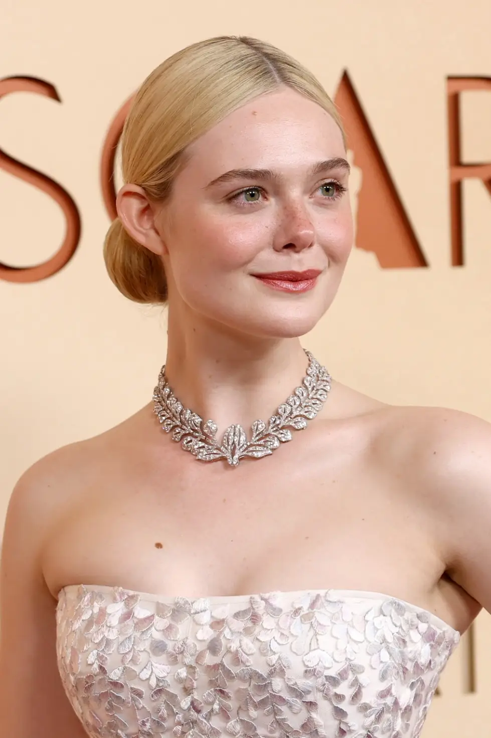 Elle Fanning, sarı saçları arkadan topuz yapılmış, ten rengi ve çilli yüzünde hafif bir gülümsemeyle, Oscar yazısının önünde, straplez, gümüşi yaprak işlemeli beyaz elbisesi ve göz alıcı yaprak desenli pırlanta kolyesiyle dikkat çekiyor.