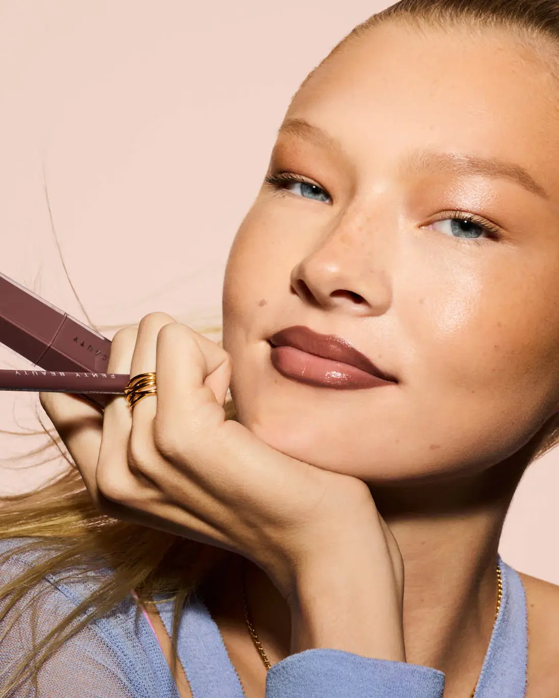 Mavi gözlü model, güneşten ısınmış bronz ve ışıltılı cildi, belirginleşen çilleri ve buğulu kahve tonlarındaki göz makyajıyla Aussie Girl estetiğinin zahmetsiz çekiciliğini yansıtırken, Fenty Beauty dudak ürünleriyle doğal parlak dudaklarını vurguluyor.