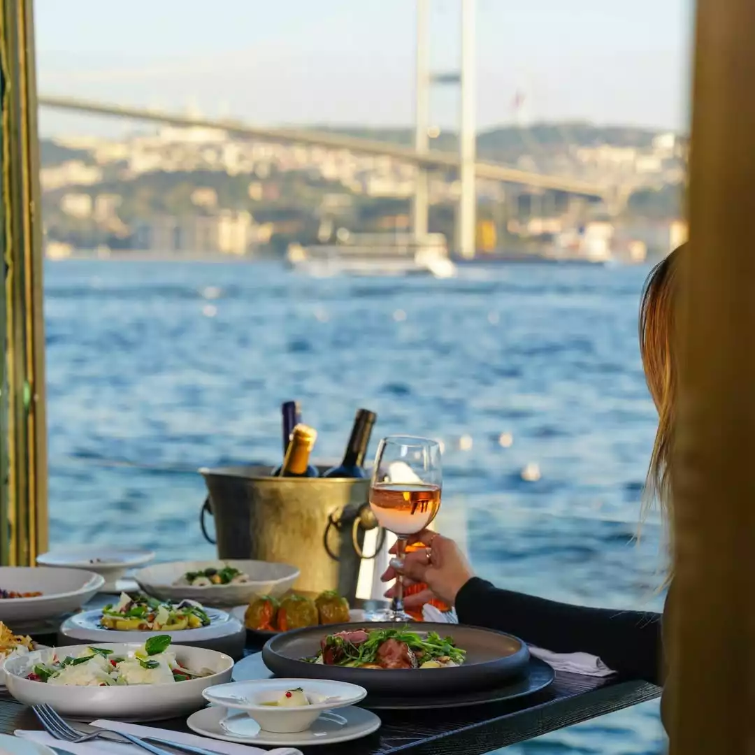 Alp Çekici –: Oggusto: Alp Çekici – Şeflerin Lezzet Rotaları. Şefin romantik buluşmalar için önerdiği Feriye Restaurant'ta Boğaz'a nazır kurulan şık sofra. Arka planda Boğaziçi Köprüsü manzarası uzanırken, elinde roze şarap kadehi tutan bir kadın özenle sunulan meze ve yemeklerin tadını
