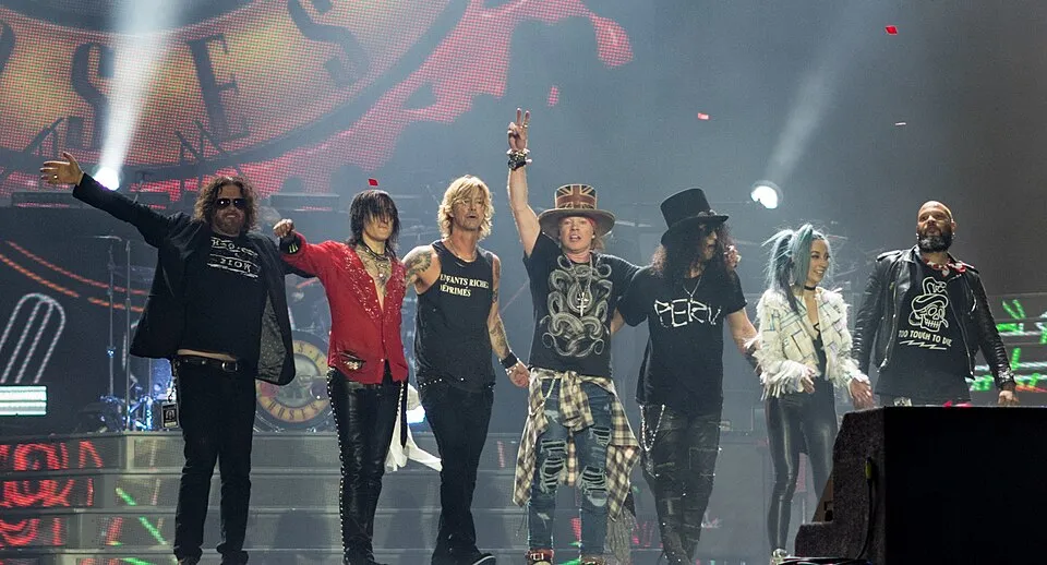 Guns N' Roses üyeleri, Axl Rose'un Union Jack şapkalı ve zafer işaretli pozu, ikonik top şapkalı Slash ve diğer grup arkadaşlarıyla birlikte kalabalığı selamlarken, arkalarındaki dev ekranda grup logosu ve havada süzülen kırmızı konfetilerle dolu sahne, 2017 London Stadium konserinden unutulmaz ve enerjik bir anı yansıtıyor.