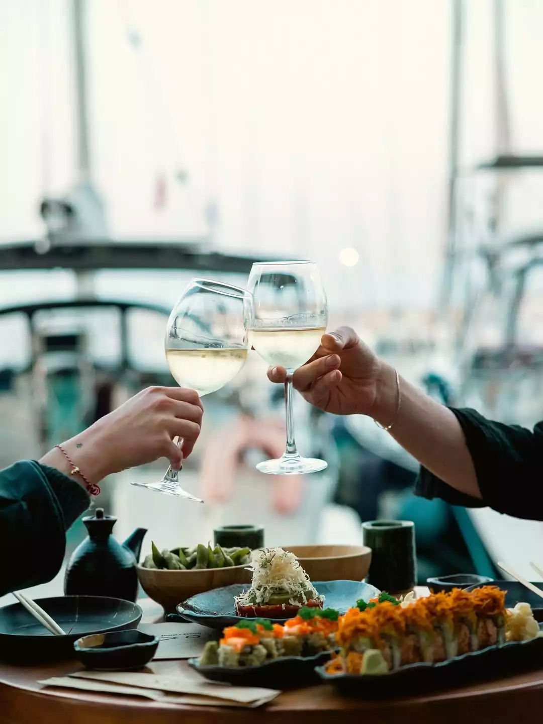 Oggusto | Salvatore Cannetiello – Şeflerin Lezzet Rotaları: Şefin romantik buluşmalar için önerdiği İoki Kandilli restoranında iki kişi, arka planda flulaşmış marina ve tekneler eşliğinde beyaz şarap kadehlerini tokuşturuyor. Masada özenle sunulmuş renkli sushi tabakları ve edamame bulunuyor.