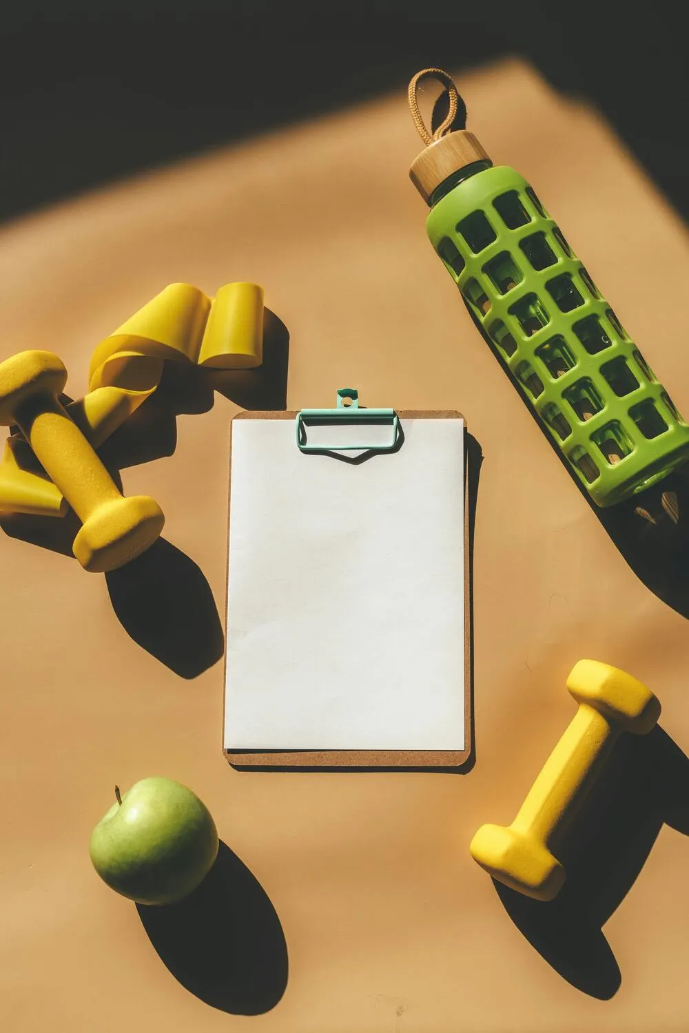 Açık kahverengi bir zeminde düz yatay çekimde, sarı dambıllar ve direnç bandı gibi spor malzemeleri, yeşil bir su şişesi, boş bir not panosu ve taze bir elma, kilo verirken kas kaybını önlemeye yardımcı olacak dengeli bir yaşam tarzının temel unsurlarını yansıtır.
