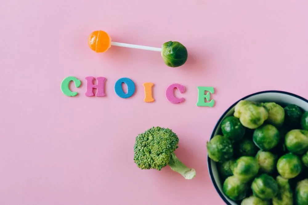 Pembe bir arka planda renkli CHOICE harfleri, bir lolipop ve brüksel lahanasından yapılmış halter figürüyle, yanında duran brokoli ve kase dolusu brüksel lahanaları eşliğinde, kilo verirken kas kaybını önlemek adına sağlıklı beslenme seçimlerinin kritik rolünü vurguluyor.