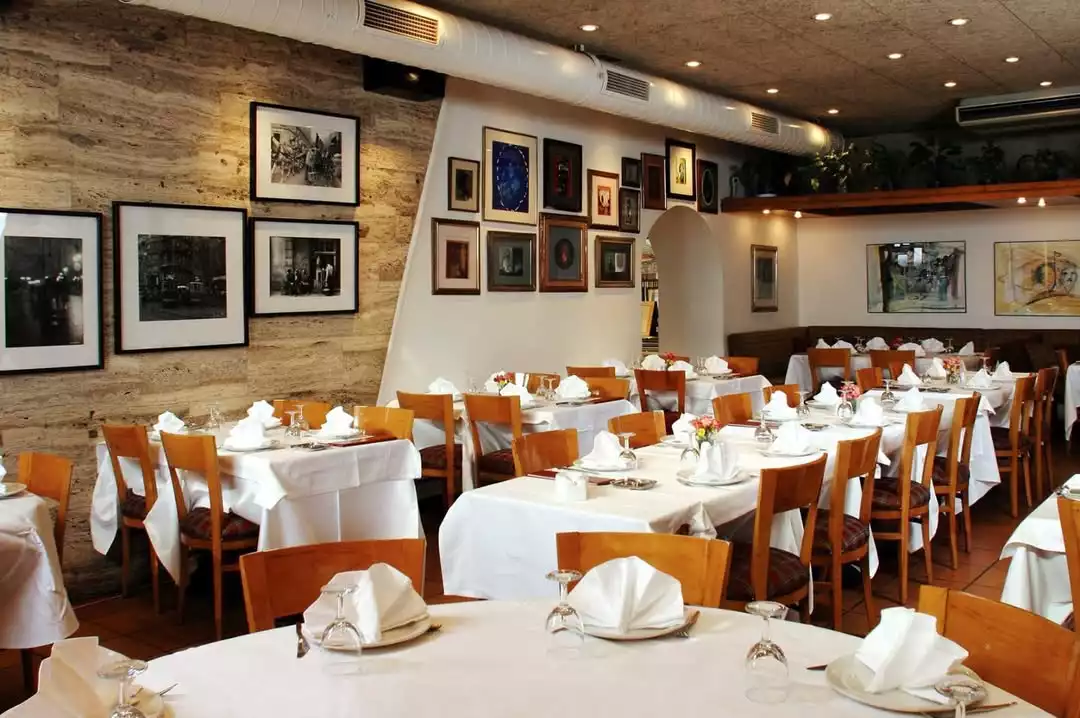 Oggusto - Salvatore Cannetiello – Şeflerin Lezzet Rotaları: Şefin balık ve meze favorisi Tarabya'daki Kıyı Balık restoranının iç mekanı. Beyaz masa örtüleri, ahşap sandalyeler, özenle katlanmış peçetelerle kurulan masalar ve taş duvarları süsleyen çerçeveli fotoğraflar sıcak bir ortam sunuyor.