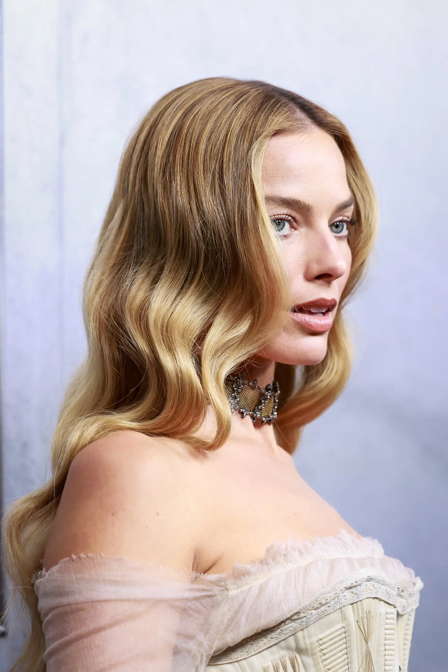 Margot Robbie, Wuthering Heights estetiğini anımsatan, dalgalı sarı saçları, dantel detaylı krem rengi korse tarzı açık omuzlu bluzu ve boynundaki zarif siyah gerdanlığıyla bu yakın çekim portrede yandan bakarak zarif bir ifadeyle poz veriyor.
